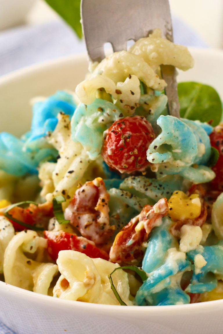 Fork lifting a bite of creamy patriotic pasta salad with red cherry tomatoes, blue rotini pasta, sweet corn, bacon, and bleu cheese dressing, perfect for 4th of July summer BBQs.