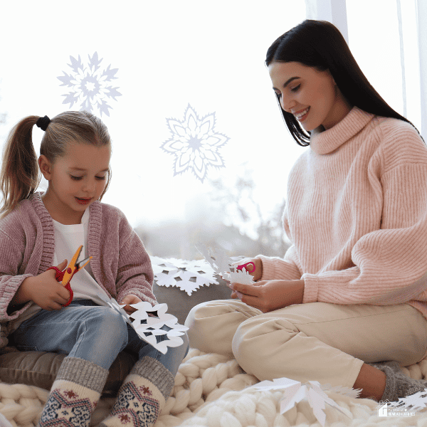 Mother and Daughter Making Paper Snowflakes near Window at Home