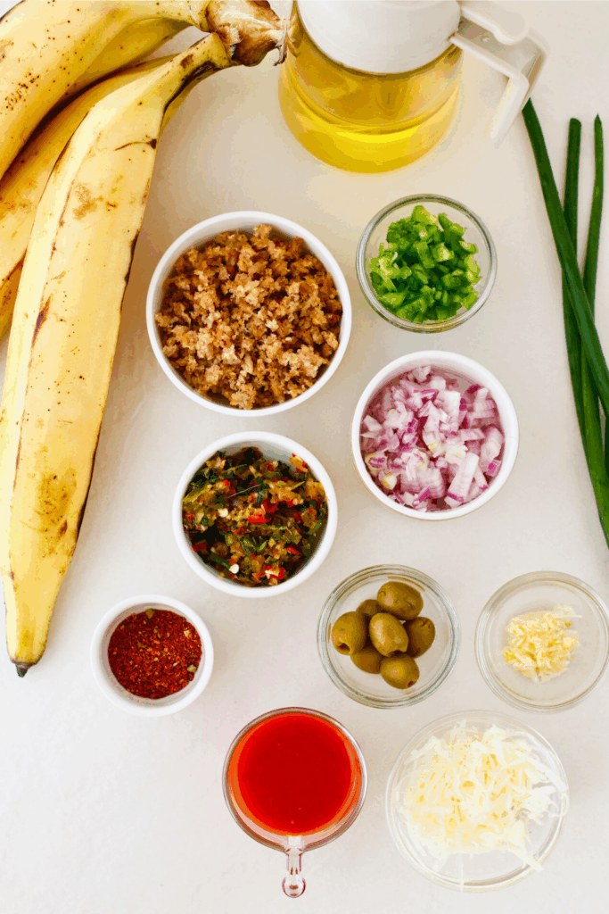 Ingredients for stuffed baked plantains (canoas de plátanos maduros), including ripe plantains, ground beef, sofrito, onion, green pepper, olives, tomato sauce, cheese, and oil laid out on a surface