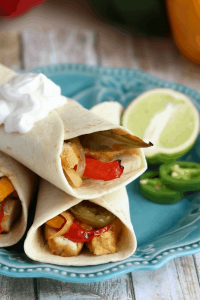 Sous Vide Tequila-Lime Chicken Fajitas wrapped in tortillas with sautéed peppers and onions, topped with sour cream on a turquoise plate.