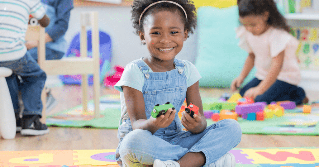 An adorable preschool age girl sits on the floor of her day care classroom and smiles for the camera. She is sitting beside a toy train track and holding two wooden train cars.