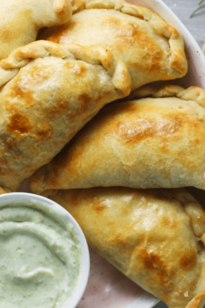 Baked Argentinian cheese and mushroom empanadas with golden flaky crust served on a plate next to a creamy green dipping sauce.