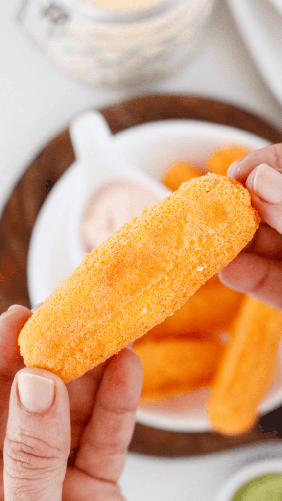 Close-up of a crispy golden sorullo held between two hands with more Puerto Rican cornmeal fritters and dipping sauce in the background.