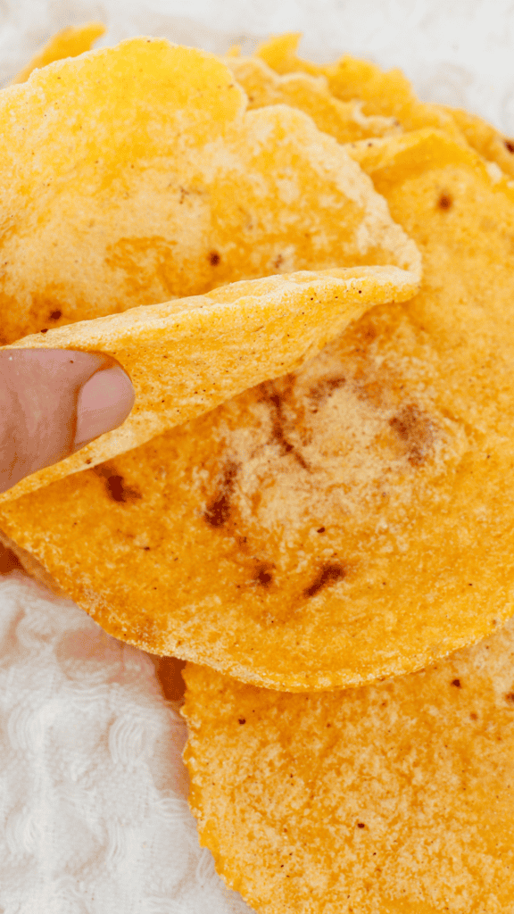 A hand gently lifts the edge of a freshly cooked corn tortilla from a stack, showing its soft texture and lightly browned surface.