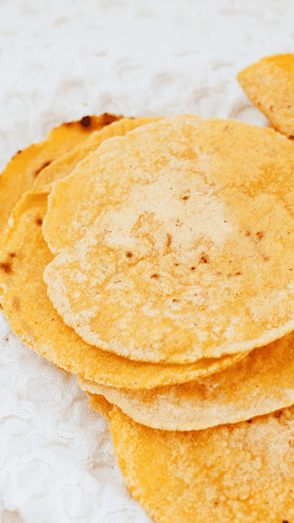 A stack of golden corn tortillas rests on a textured white cloth, showing their rustic shape and lightly toasted spots.