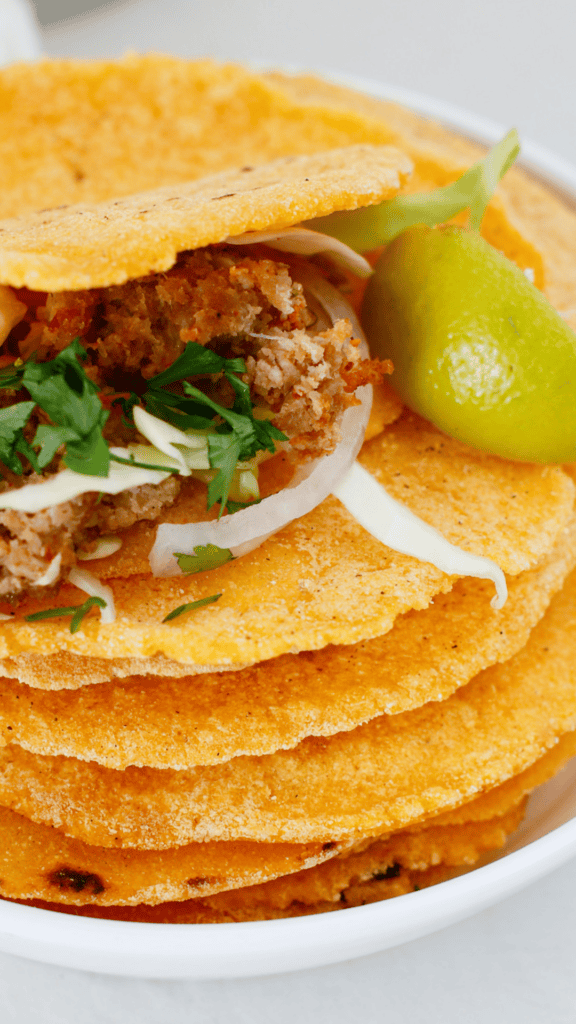 A homemade corn tortilla is filled with seasoned meat, shredded cabbage, onion, and cilantro, with a fresh lime wedge beside it.