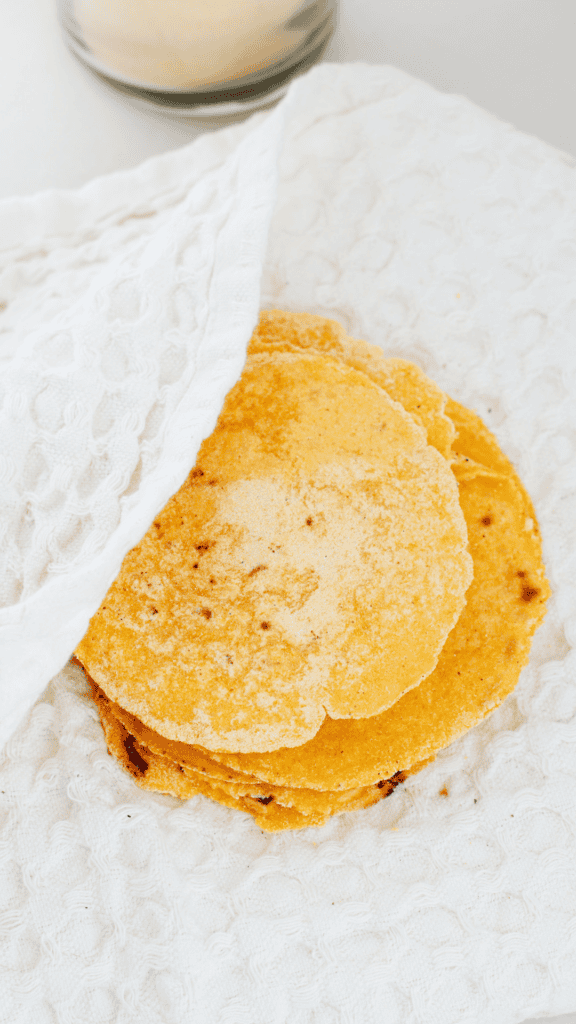 A stack of freshly cooked corn tortillas is tucked inside a textured white kitchen towel to help keep them warm and soft.