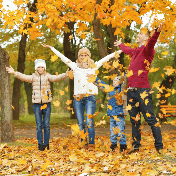 A family plays together under colorful autumn trees as falling leaves surround them during a fun outdoor fall activity.