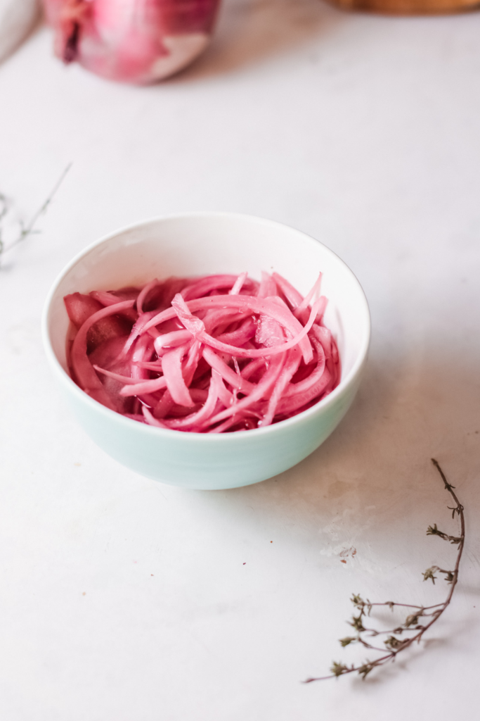 Small white bowl filled with homemade quick pickled red onions on a clean kitchen counter with a whole red onion in the background.