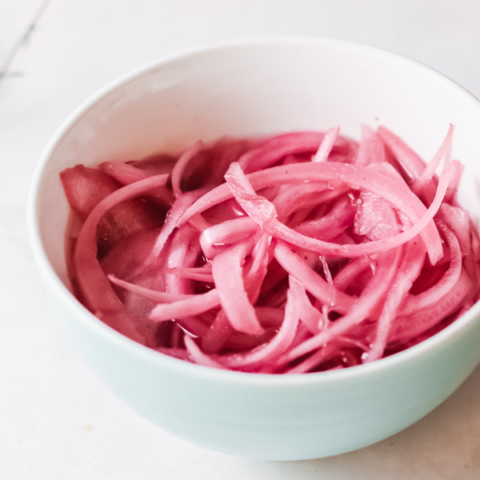 Bowl of bright pink quick pickled red onions in brine on a light marble surface.
