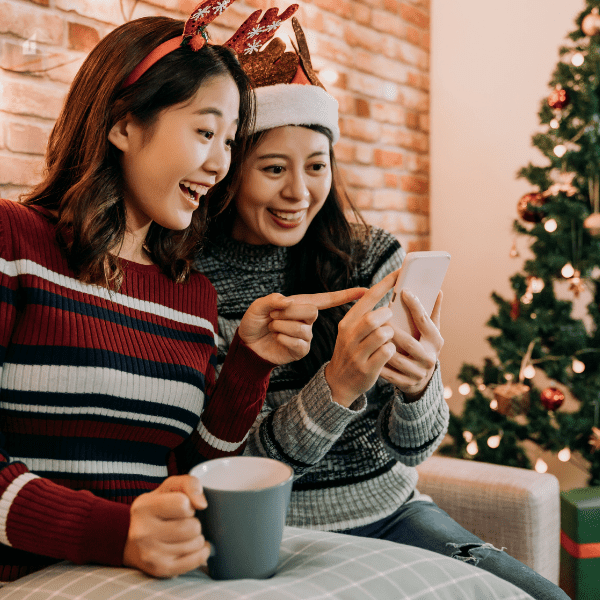 Two women wearing Christmas accessories excitedly look at a phone while sitting near a decorated tree, symbolizing holiday shopping and online budgeting during the Christmas season.