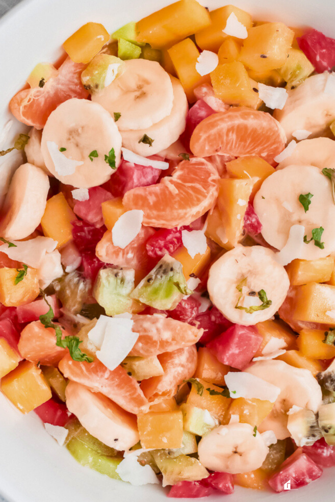 Close-up of Puerto Rican fruit salad featuring banana, kiwi, mango, tangerine, and strawberries, mixed with sweetened condensed milk and topped with coconut flakes.