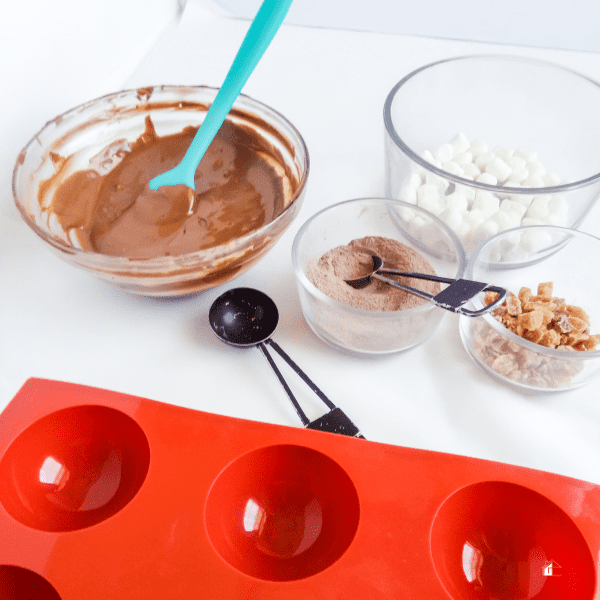 ingredients to make Ginger Dark Chocolate Hot Cocoa Bombs. mold, ginger bites, melted chocolate, mini marshmallow in glass bowl, powder and a scoop