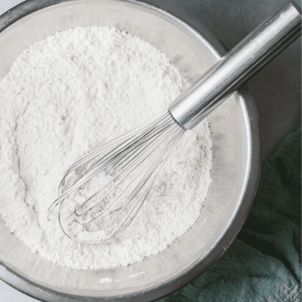 dried ingredients such as flour, sugar and baking powder in a mixing bowl with a whisk over it.