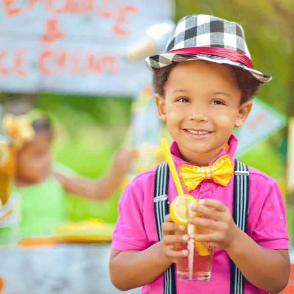 Little boy holding a lemonade in front of a lemonade stand.
