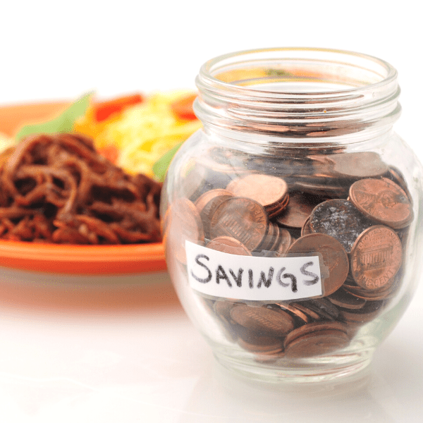 A glass jar holding pennies inside labeled savings. Behinds it a plate of food.