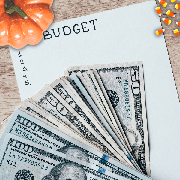 Cash on top of a paper with the text budget and 5 numberic bullet points. On the left there's a Halloween pumpkin and candy corn.