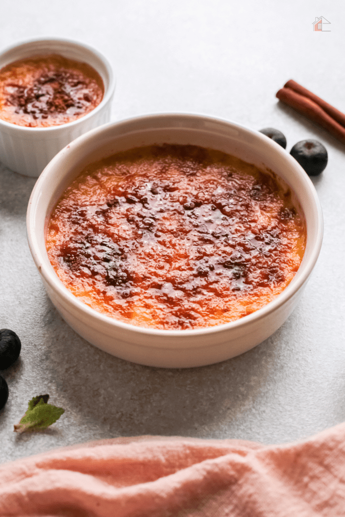 Crema catalana serves with smaller serving behind it and blue betties and cinnamon sticks in the background.