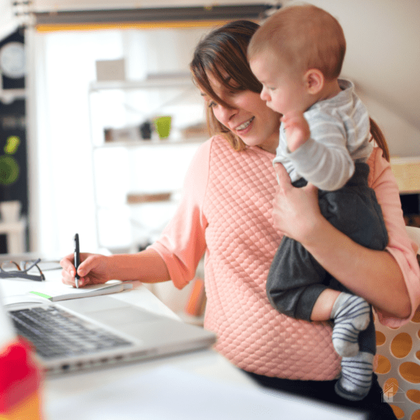 Mom holding baby while writing something on her note bad and looking at her laptop.