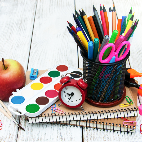 Assorted school supplies including colored pencils, scissors, notebooks, paint set, and an apple arranged on a table for budget-friendly school preparation.