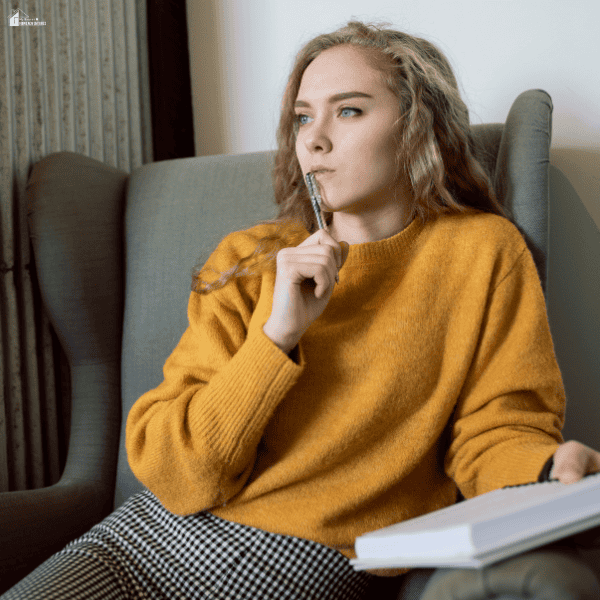 A woman sitting in a chair with a notebook and pen while thinking carefully about her budget, expenses, and spending priorities.