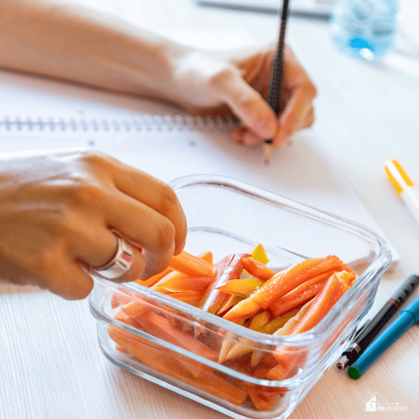 An image of hands, holding a pencil and eating vegetable sticks on the other.