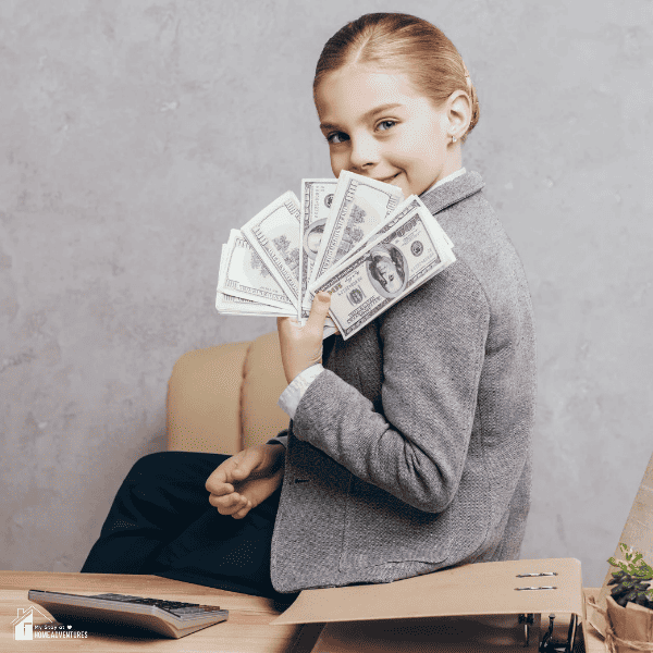 portrait of smiling little girl showing money in hand while sitting on table isolated on grey