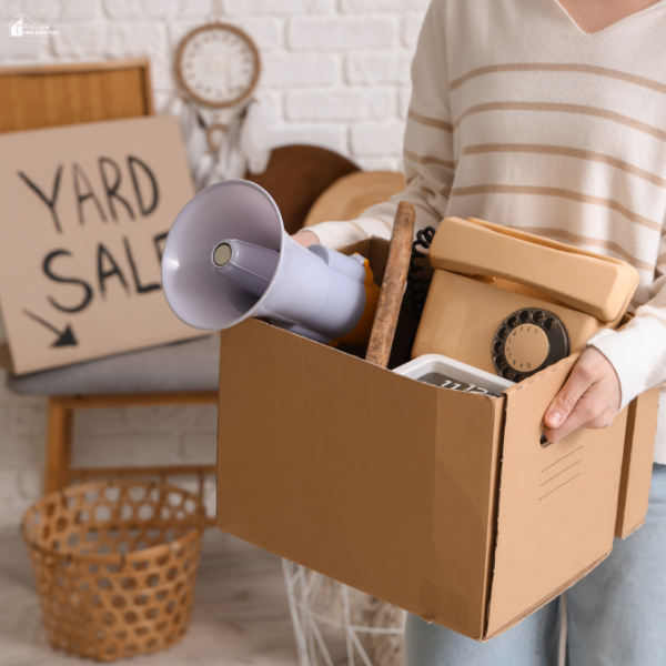 A person holding a cardboard box filled with vintage items like a rotary phone and megaphone, preparing for a yard sale.