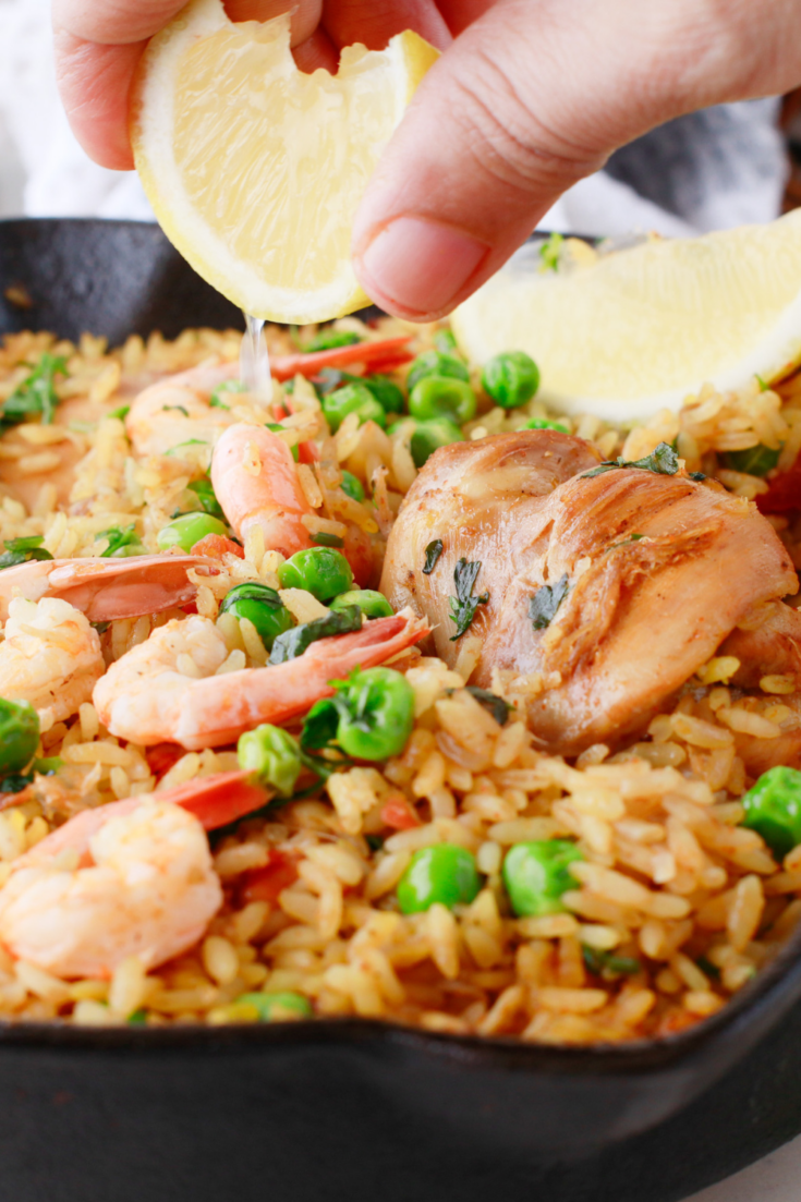 A close-up of an easy chicken and shrimp paella in a cast iron skillet, featuring saffron rice, green peas, juicy chicken thighs, shrimp, and a lemon wedge being squeezed on top.
