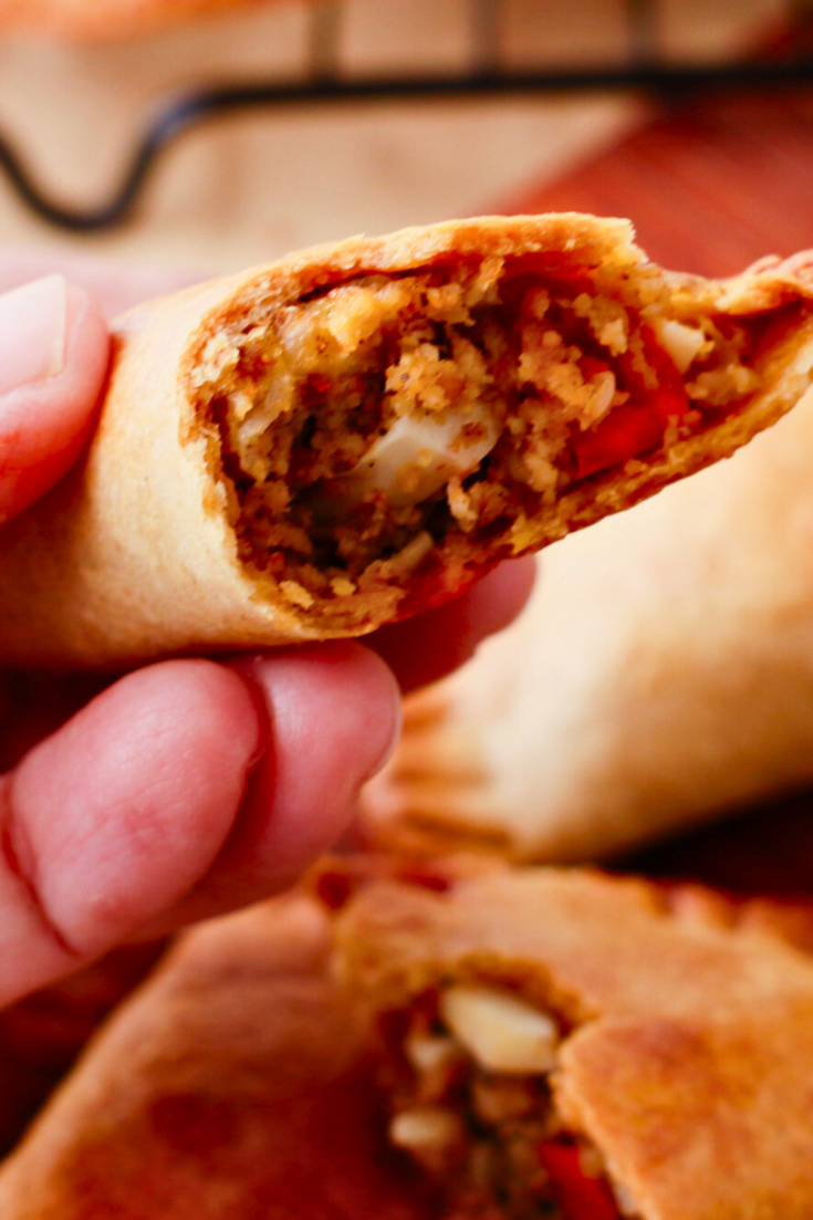 Hand holding a flaky baked beef empanada with a bite taken out, showing the savory ground beef, red pepper, onion, and egg filling inside the crispy crust.