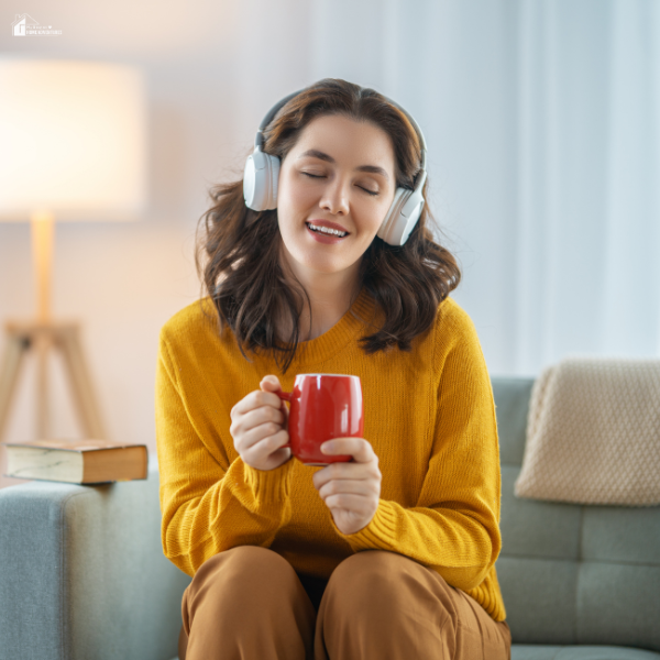 A woman sits on a couch wearing headphones, smiling and enjoying a cup of coffee.