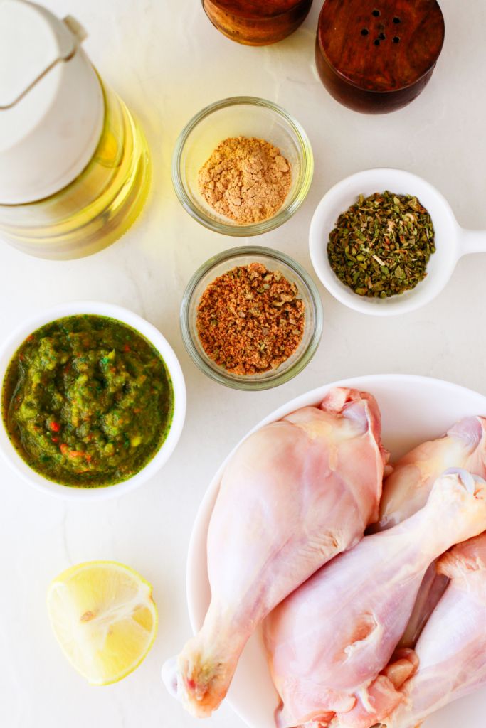 Raw chicken drumsticks, sofrito, adobo seasoning, garlic powder, oregano, olive oil, and lime displayed together as fresh ingredients for Puerto Rican fried chicken.