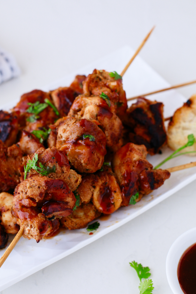 Grilled Puerto Rican pinchos made with juicy BBQ chicken skewers, topped with sauce and herbs, served on a white plate with bread on the side.