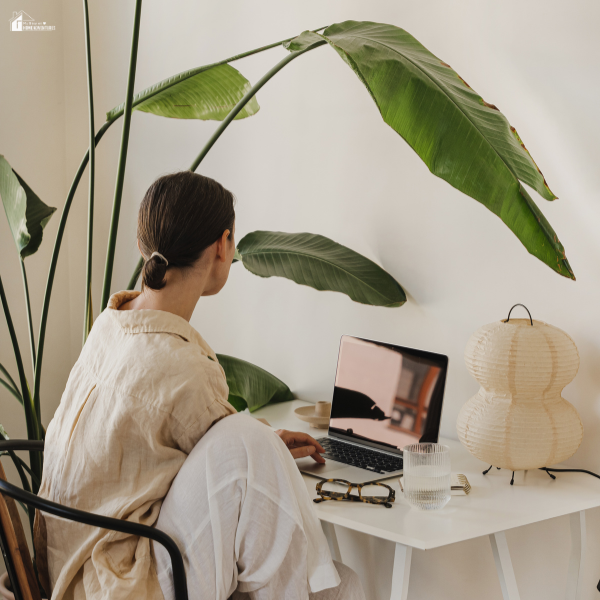 A woman working on a laptop at a small white desk with a large leafy plant in the background and soft ambient lighting.