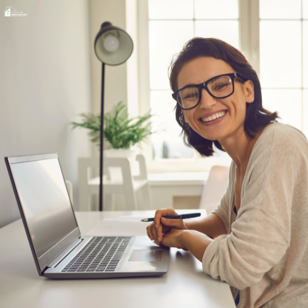 A smiling woman sits at a desk with a laptop, pen in hand, in a bright home office space.