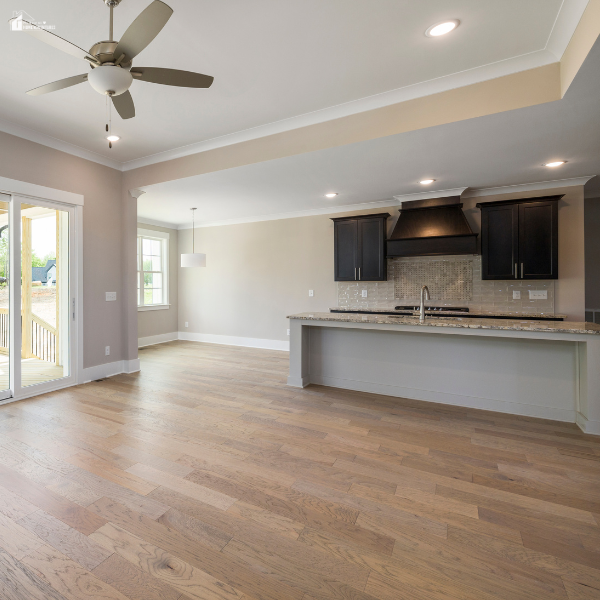 An empty open-concept kitchen and living space features clean, modern hardwood flooring and large windows with natural light.