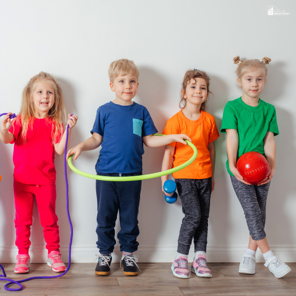 Four young children in colorful outfits smile while holding a jump rope, hula hoop, dumbbell, and red ball against a white wall in a homeschool.