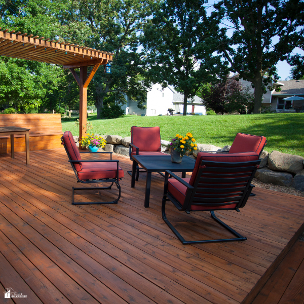 A backyard composite deck with red cushioned patio chairs, a table, and a pergola, set against a green lawn and trees.