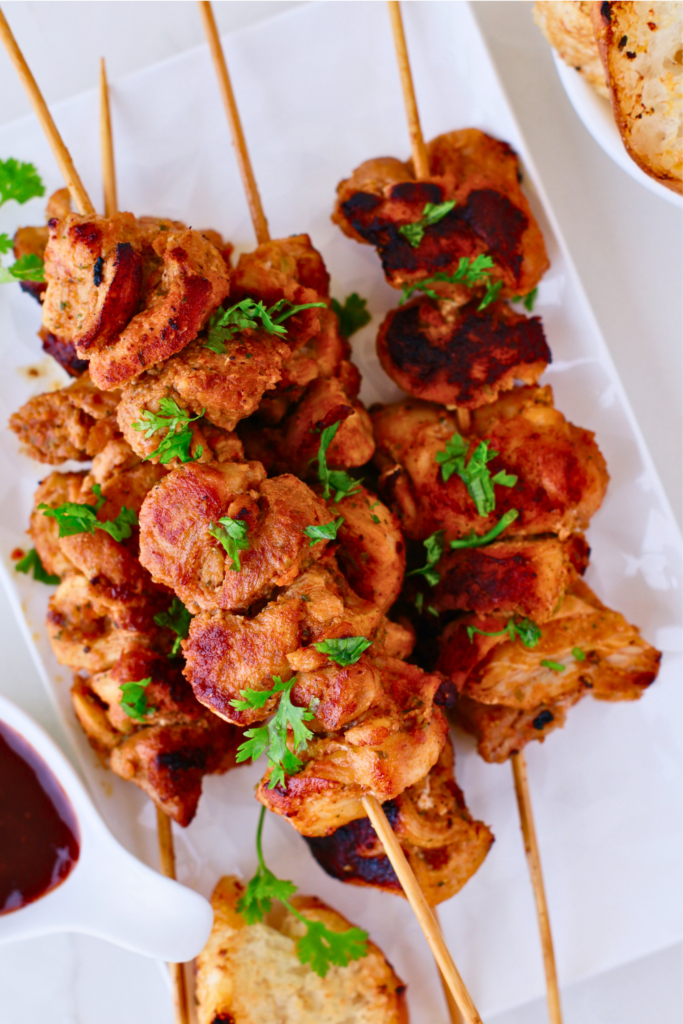 Grilled Puerto Rican pinchos served on skewers, garnished with fresh herbs, paired with bread and BBQ sauce, showcasing an easy pinchos recipe.