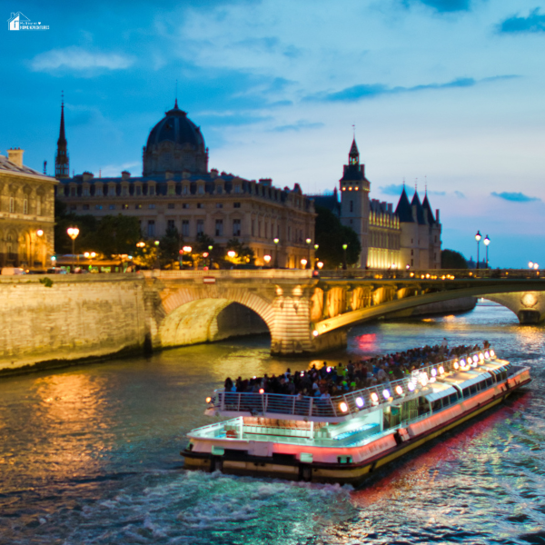 A river cruise boat glides under a lit stone bridge at dusk, surrounded by historic European buildings and glowing city lights.