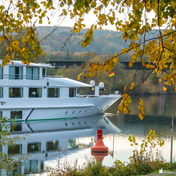 A modern white cruise ship floats on a peaceful river framed by colorful autumn leaves and gentle hills in the background.
