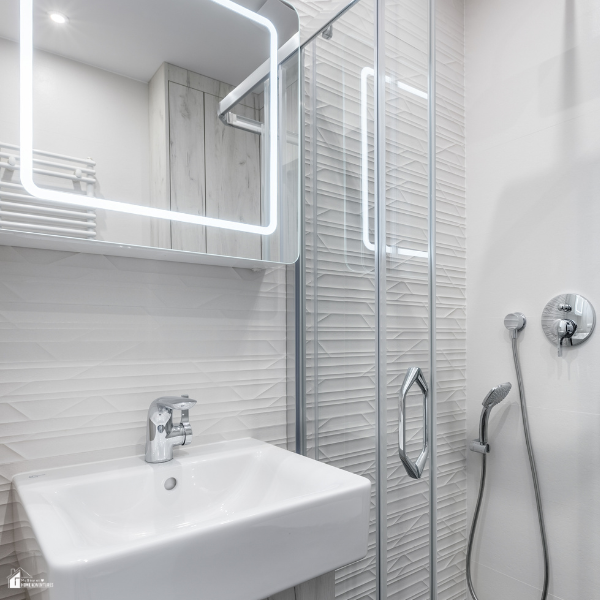 A modern bathroom with a bright LED-lit mirror above a white sink and a glass shower enclosure beside it.