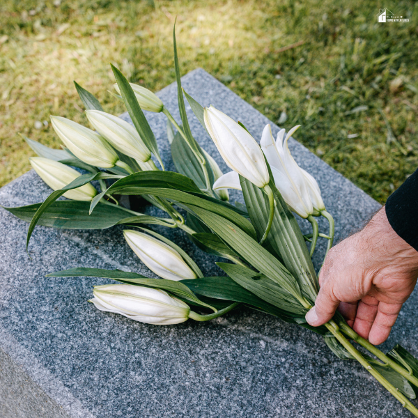 A hand placing a bouquet of white lilies on a gray gravestone in a peaceful grassy cemetery.