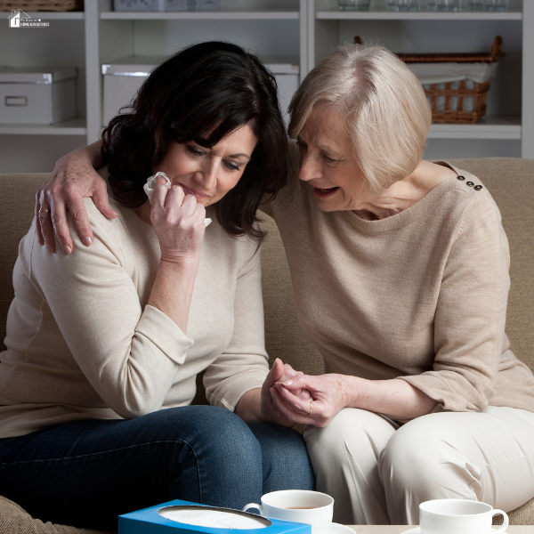 An elderly woman comforting a younger woman who is crying while holding a tissue, both seated on a couch.