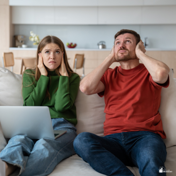 A couple sits on a couch covering their ears in frustration, reacting to loud noise in their apartment.