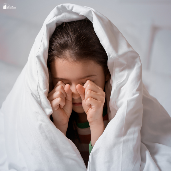 A young child wrapped in a white blanket sits on a bed, rubbing their eyes as they wake up from a disrupted sleep.