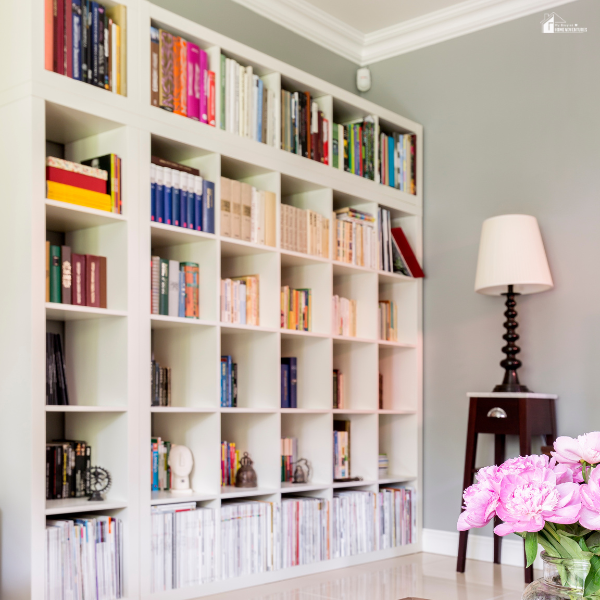 A modern living room features a large white bookshelf filled with colorful books and decor, next to a tall lamp and pink flowers in the foreground.
