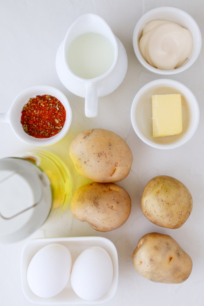 Overhead view of ingredients for Puerto Rican mashed potatoes with eggs, including potatoes, eggs, mayonnaise, butter, milk, olive oil, and adobo seasoning—essential items for making this creamy Caribbean side dish.