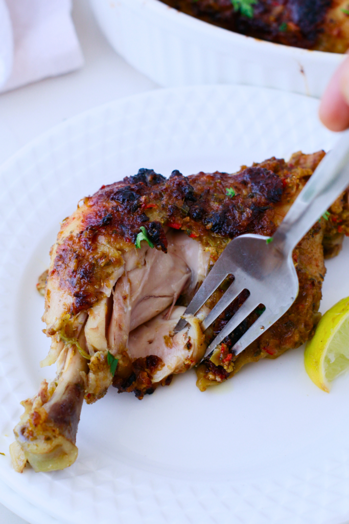 Close-up of tender Puerto Rican baked chicken with crispy skin and juicy meat, being cut with a fork on a white plate, served with a fresh lime wedge.