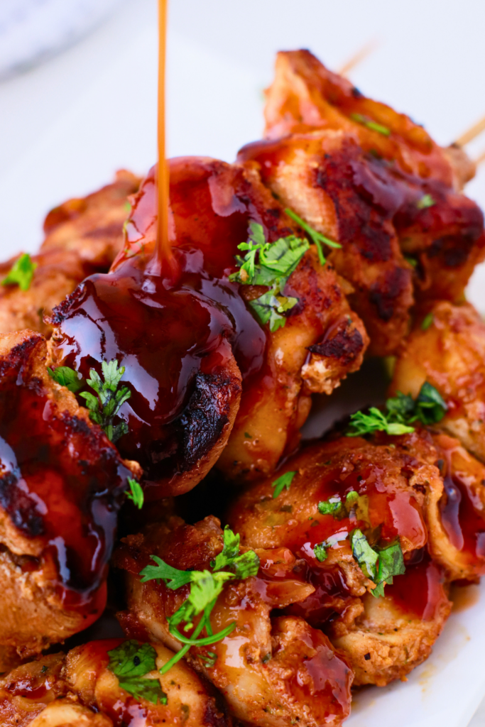 Close-up of Puerto Rican chicken pinchos with BBQ sauce being drizzled over grilled meat, garnished with fresh cilantro on a white plate.